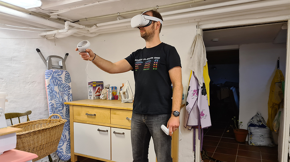 man med VR-headset och kontroller står i ett källarrum med tvättutrustning. Foto. 
