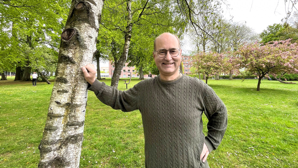 En glad man står lutad mot ett träd utomhus. Foto. 