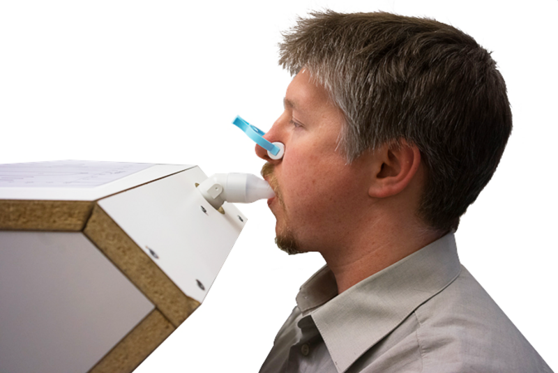 Person med klämma för näsan blåser i ett rör. Foto. 