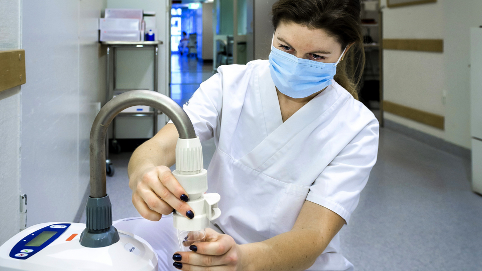 kvinna i ansiktsmask hanterar mätutrustning i en sjukhuskorridor. Foto. 