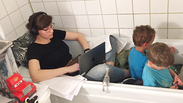kvinna sitter i badkaret med dator och arbetar. två barn sitter i fotänden. Foto. 