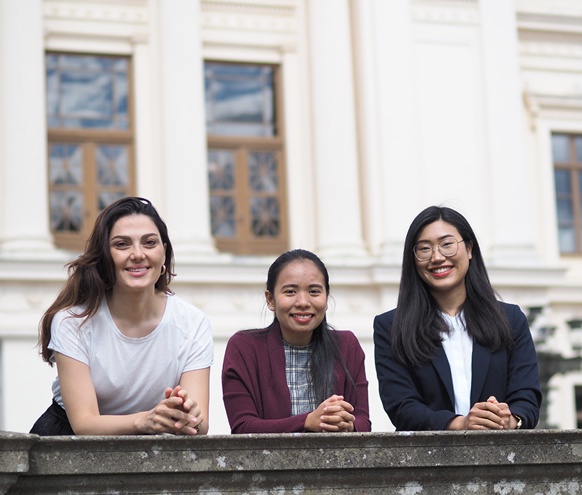 Tre studenter framför en vit byggnad. Foto. 