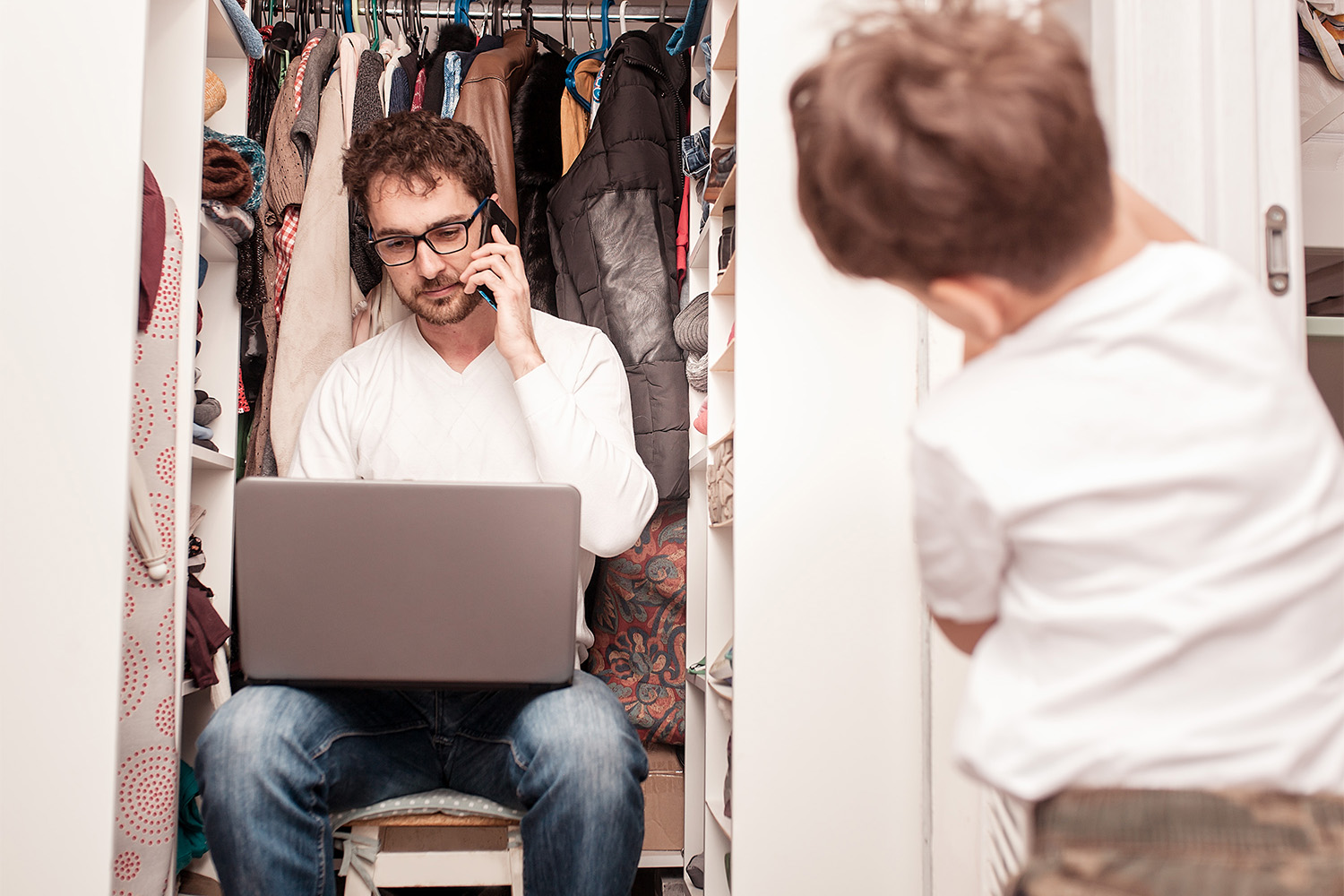 en man sitter i klädkammaren med laptop och telefon, ett barn kikar in. Foto. 