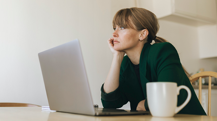 person sitter vid köksbordet med laptop och ser uttråkad ut. Foto. 