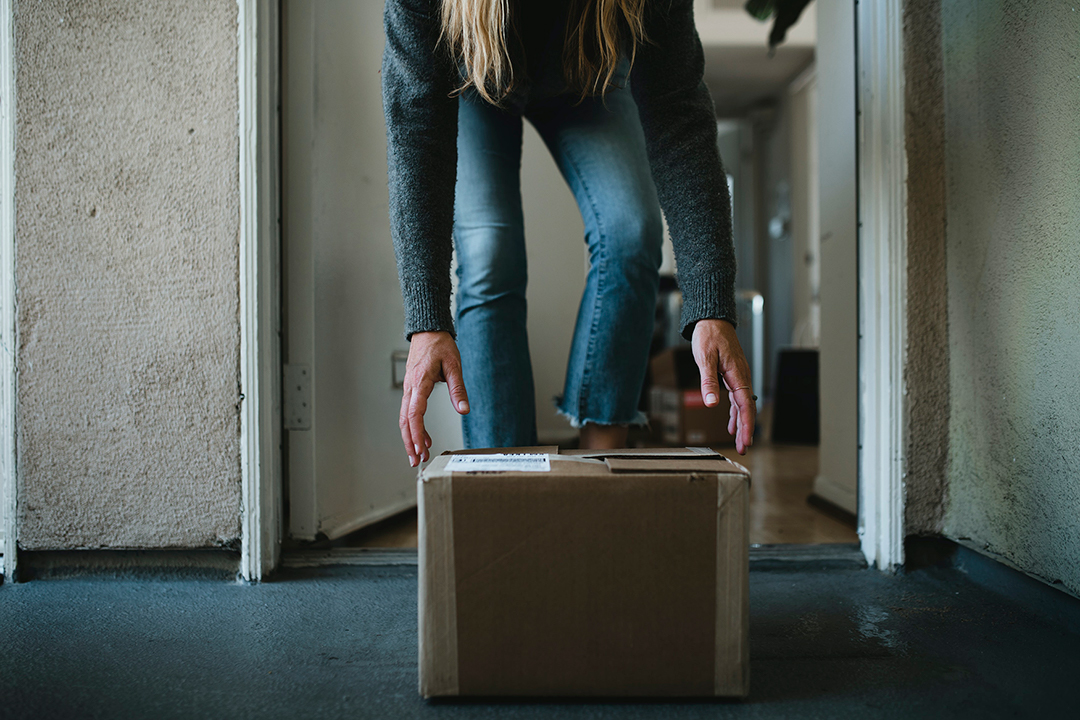 person böjer sig ner till ett paket utanför en dörr. Foto. 