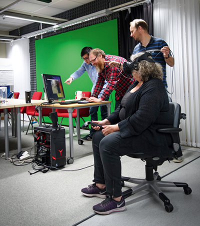 a person with a VR-headset is sitting on a chair while three people work with a computer in the background. Photo.