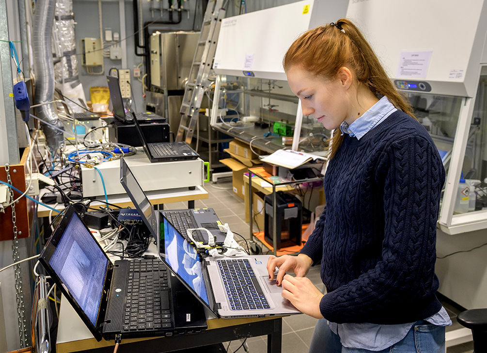 person står i ett laboratorie omgiven av datorer och utrustning. Foto. 