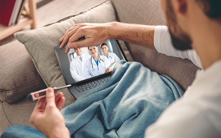 en person tar tempen på en laptop som ligger nedbäddad i en soffa. på skärmen syns tre läkare. Foto. 